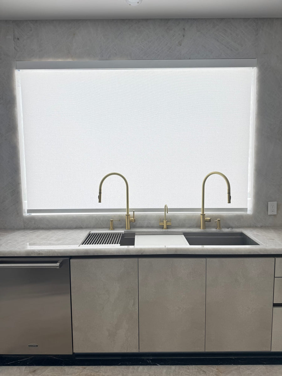 Horizontal kitchen window with a Designer Roller shade above a double sink and two brass faucets